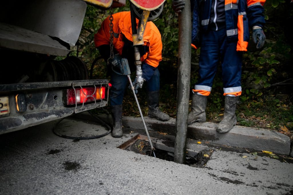 Male workers cleaning storm drains from debris using specialized pump and tools.